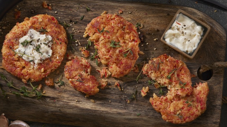 Three crab cakes, two broken up, with a small container of tartare sauce on a wooden chopping board