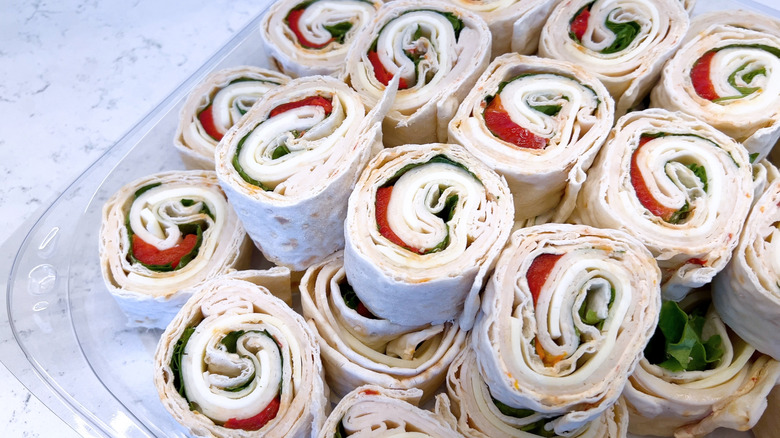 Pinwheel sandwiches with meat, cheese, tomato, and lettuce stacked in plastic container