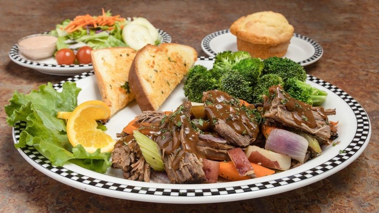 pot roast with gravy, celery, onions, carrots, and potatoes on a plate with broccoli and garlic toast, with side plates of salad and a cornbread muffin