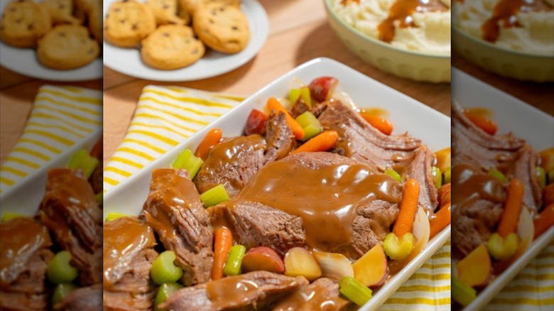 a rectangular plate of pot roast with carrots, celery, onions, sliced potatoes, and gravy next to a bowl of mashed potatoes and a plate of cookies