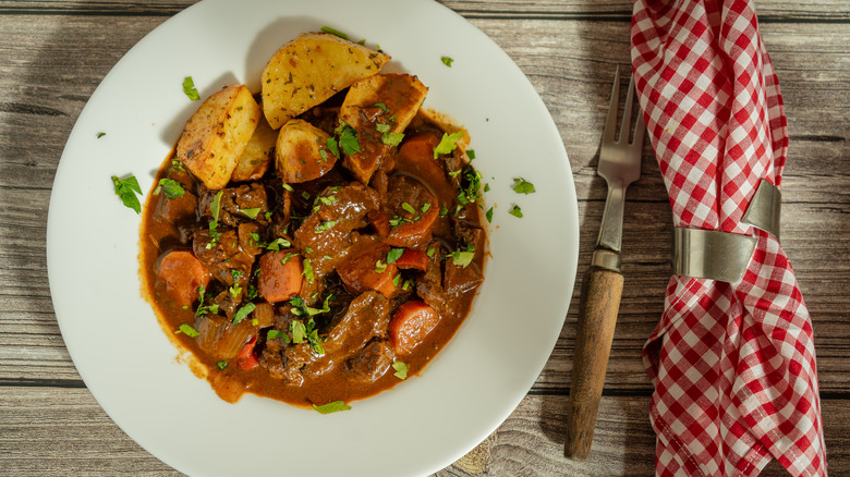 a bowl of pot roast with onions, carrots, sliced potatoes, parsley, and plenty of gravy on a wooden table