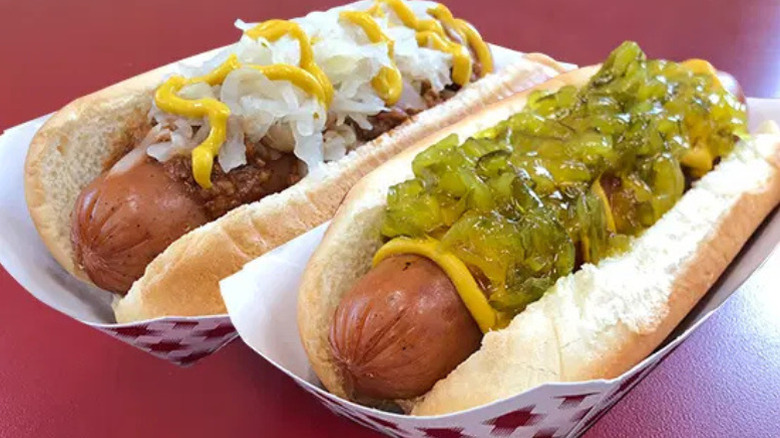 one hot dog with chili, sauerkraut, and mustard, and one hot dog with mustard and relish against a red background