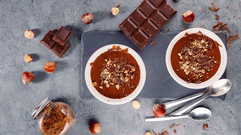 Two servings of chocolate mousse with chocolate bars and hazelnuts around them.