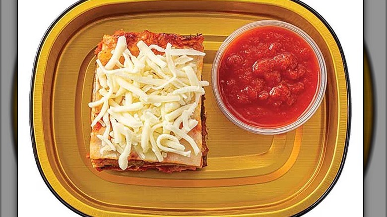 A portion of baked eggplant with shredded cheese on top and a tub of tomato sauce on the side