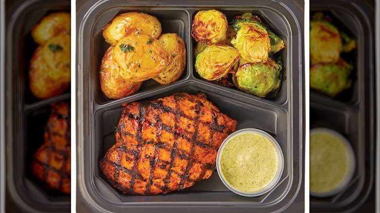 A plastic container with roasted chicken, roasted potatoes, brussels sprouts, and a tub of white sauce