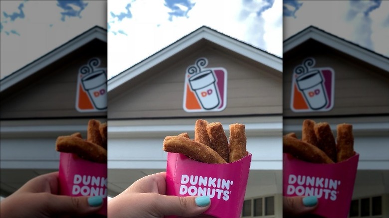 A hand holding a cardboard takeout container of Dunkin' Donut Fries in front of a storefront