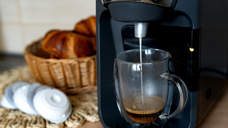 Coffee maker brewing a cup of coffee next to coffee capsules and a basket of bread