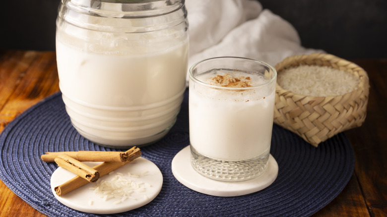 A glass of horchata on ice next to a pitcher, cinnamon sticks, and a basket of rice