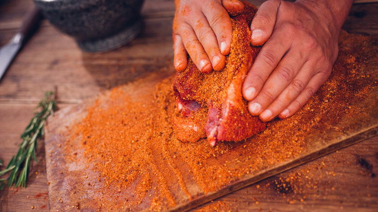 Hands press a dry rub onto a raw piece of meat on a cutting board before cooking