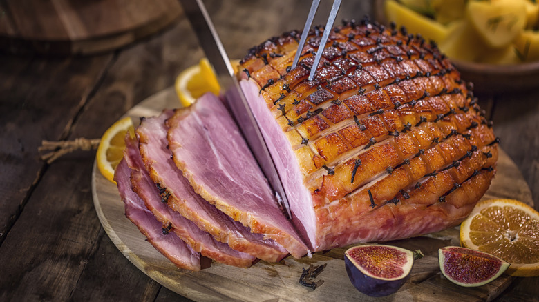 Someone carving a honey baked ham with figs and lemon slices next to it on a wooden carving board