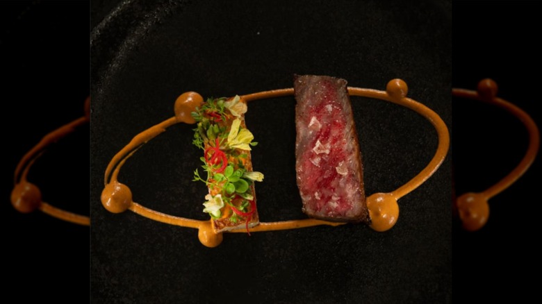 A black plate with a slice of salted meat, edible flowers, and a circle of brown sauce