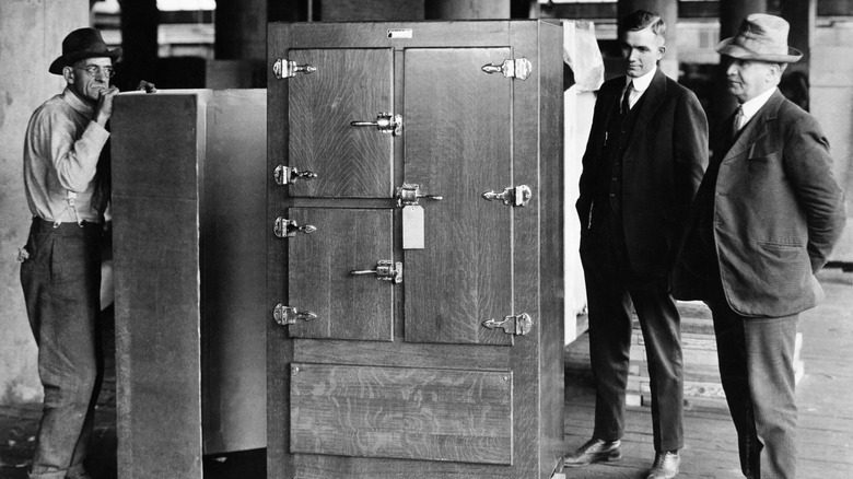 A black and white photograph of three men standing next to an early refrigerator