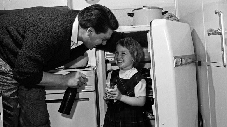 A black and white photo of a 1950s or 1960s man and child, with the man appearing to hold a beer while telling the smiling child not to hide in the open refrigerator