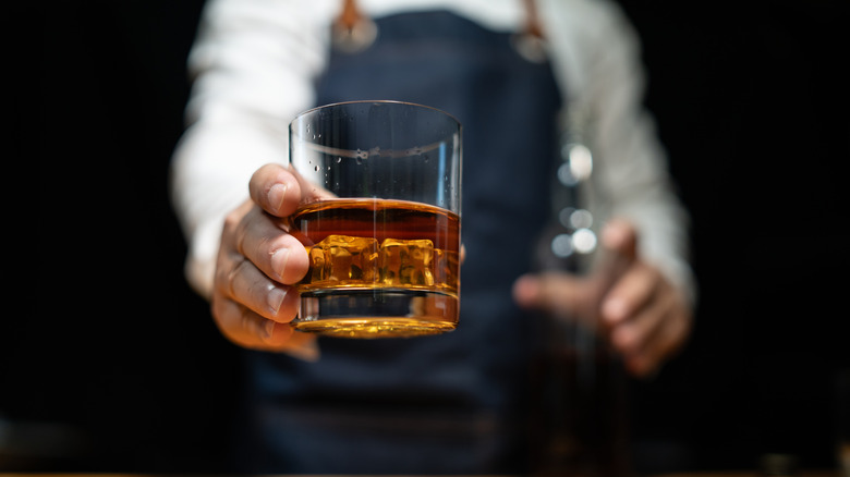 A close up of a bartender handing someone a glass of whisky on the rocks