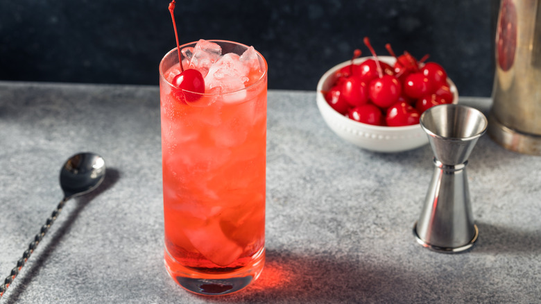 A Dirty Shirley cocktail on a bartop with a stirring spook, jigger, and bowl of cherries