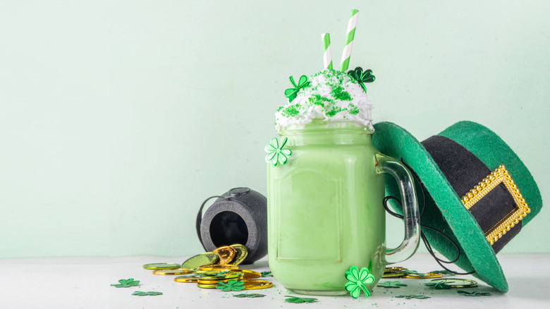 Mint milkshake in a mason jar mug with a spilled pot of gold, green hat, and other St. Patrick's Day decor