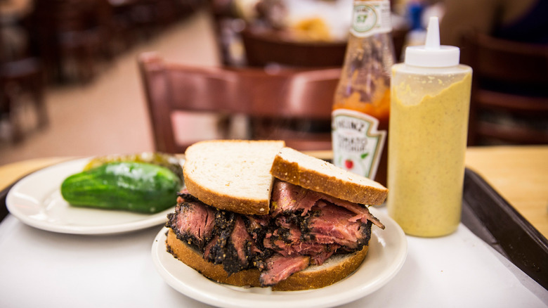 A pastrami sandwich on rye bread next to a bottle of spicy mustard, pickles in the background, at Katz's Delicatessen