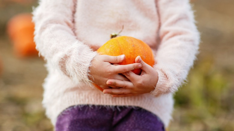 A kid holds a bright orange pumpkin against a fluffy pale pink sweater and purple pants