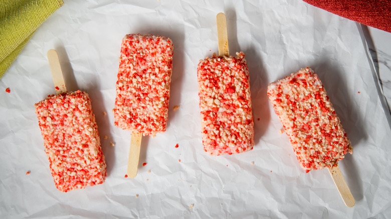 Four strawberry shortcake ice cream bars arranged on white paper with loose crumbles