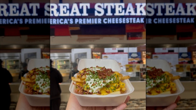 A hand holds a box of loaded fries in front of a Great Steak restaurant in a mall