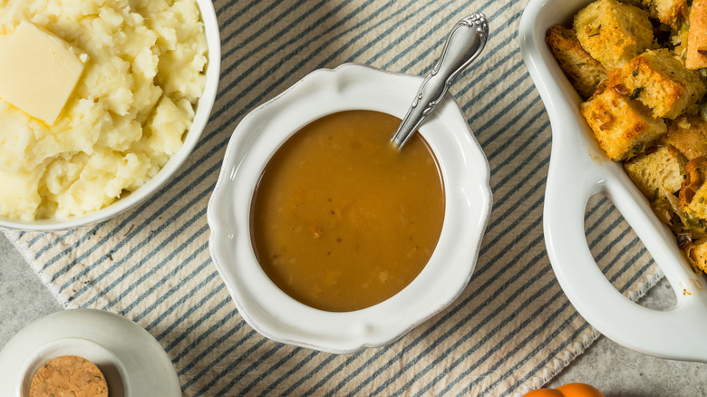 Gravy in a white bowl next to a bowl of mashed potatoes and a tray of baked stuffing