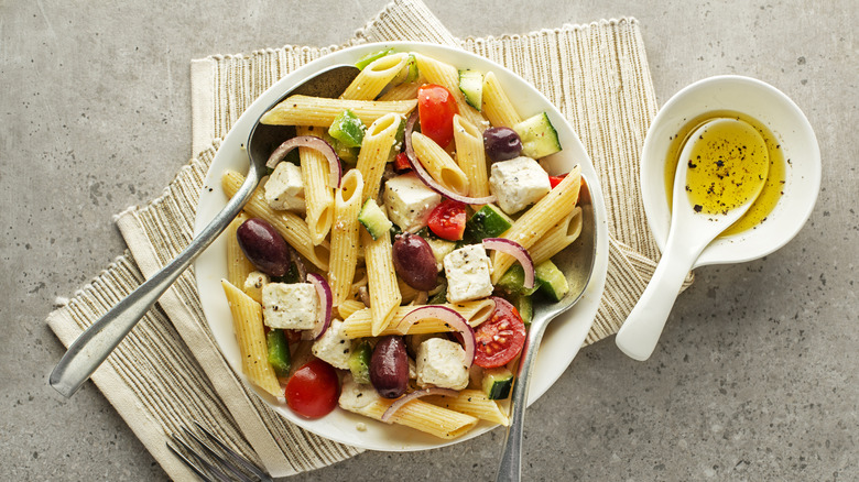 Pasta salad with olives, tomatoes, and feta cheese next to a dish of oil