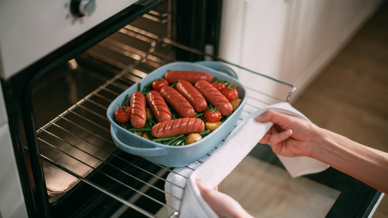 Hands take a baking dish of sausages and vegetables out of an oven
