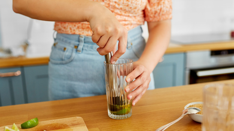 Woman in jeans muddling lime in a glass on a wooden countertop.