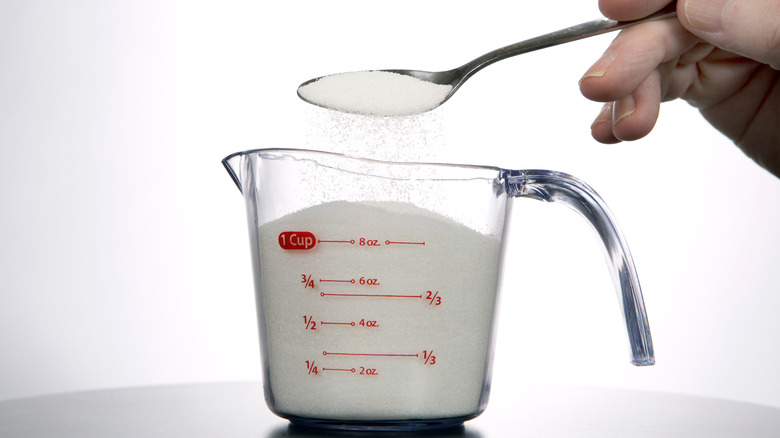 A person pours a spoonful of sugar into a glass measuring cup