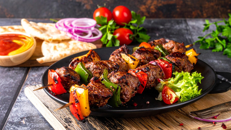 Steak kebabs with bell peppers on a black plate with pita, sauce, tomatoes, onions, and parsley in the background.