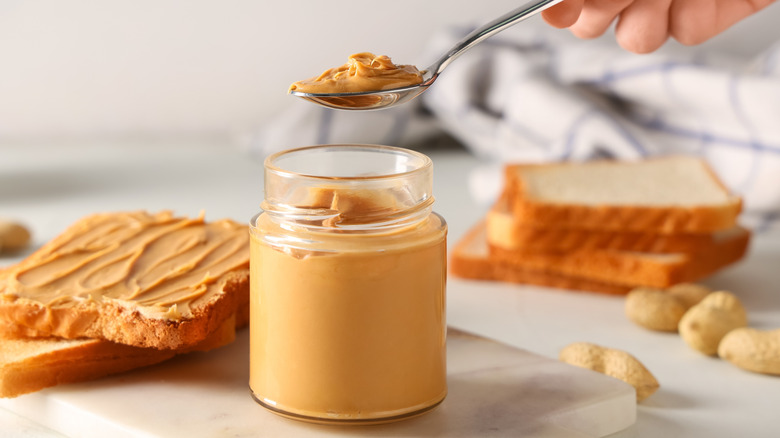 A hand holding a spoon with a scoop of homemade peanut butter taken out of a glass jar beside slices of bread and peanuts