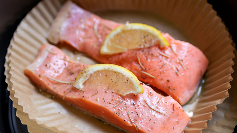 Raw salmon filets on top of a brown air fryer liner, with seasoning, lemon slices, and extra oil