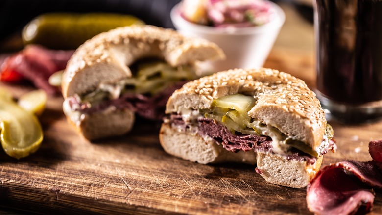A pastrami bagel with pickles, halved, on a wooden board