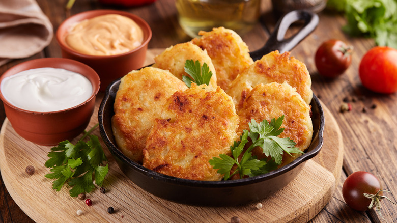 Crispy, golden hash browns in a dish next to some dipping sauce, surrounded by tomatoes and herbs