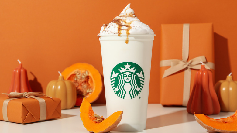A white paper cup of Starbucks' pumpkin spice latte, with pumpkin slices, gourd candles, and orange boxes