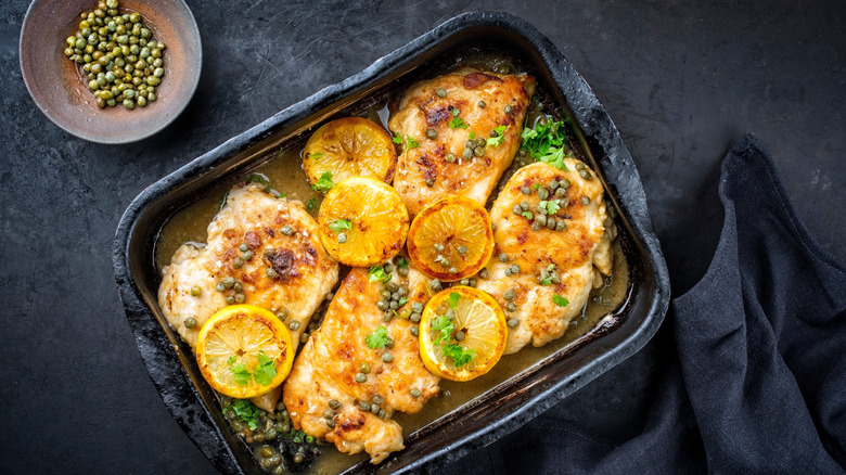 Chicken Piccata in a pan against a dark countertop with a bowl of capers on the side.