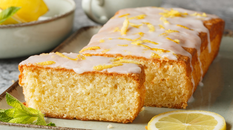 Sliced lemon pound cake with glaze and zest on top with a sliced lemon on the side.