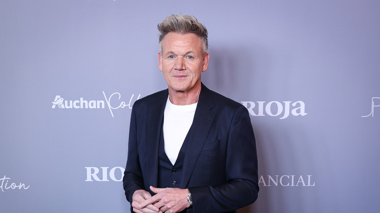 Gordon Ramsay posing for a camera at a red carpet event against a pale purple background.