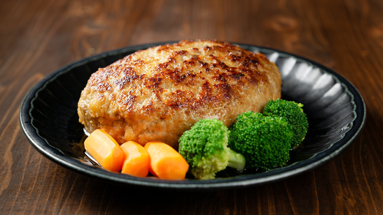 A hamburg steak on a black bowl with carrots and broccoli, on a wood table.