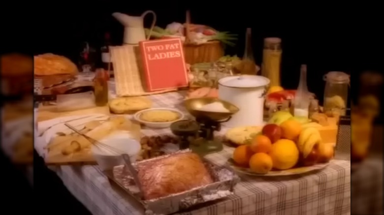 Title card for Two Fat Ladies with the show name on a red book, on a table covered in food