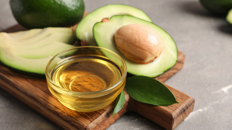 A glass bowl of avocado oil with avocadoes in the background