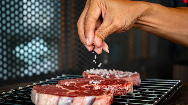 A person seasoning steaks on a grill with coarse salt