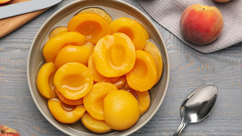 Canned peaches in a white bowl surrounded by fresh peaches