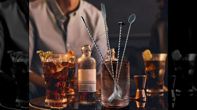 Bar setting with Jetkong bar spoons standing in a spouted glass and surrounded by drinks, a cocktail mixer, and jigger with a person blurred in the background