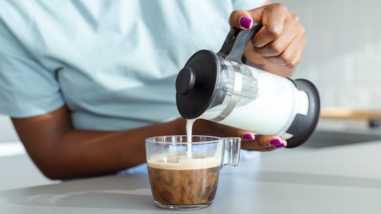 Woman pours hot milk from a French press into a cup of coffee