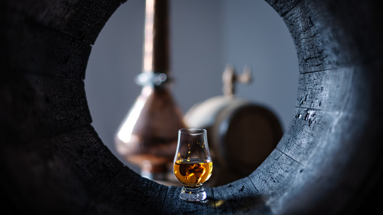 Glass of whiskey inside oak barrel next to still