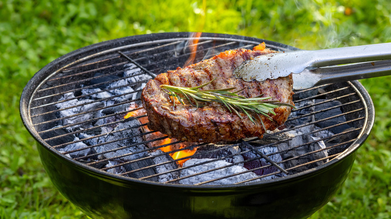 A thick steak grilling in the center of a charcoal grill on a lawn.