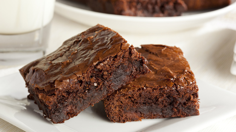 Two slices of chocolate brownies on a white plate with the edge of a full plate of brownies in the backdrop.