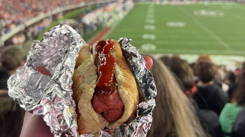 Hand holding hot dog with ketchup in foil in football stadium
