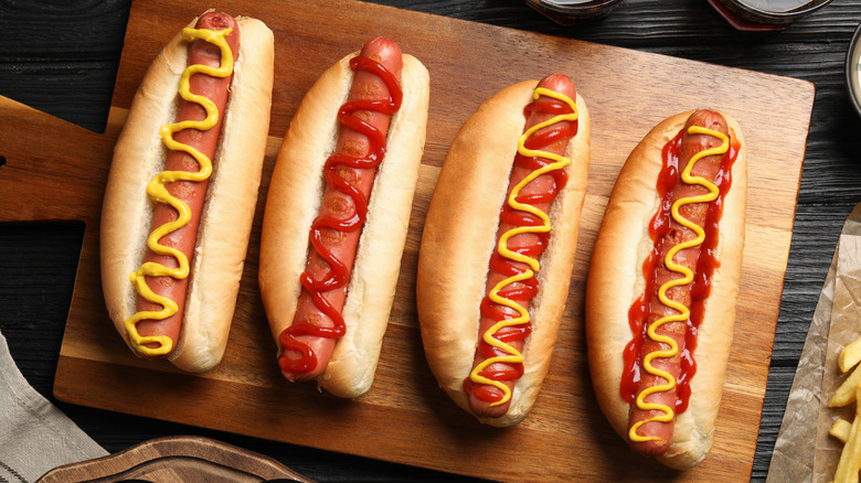 Four hot dogs with ketchup and mustard on wooden cutting board.